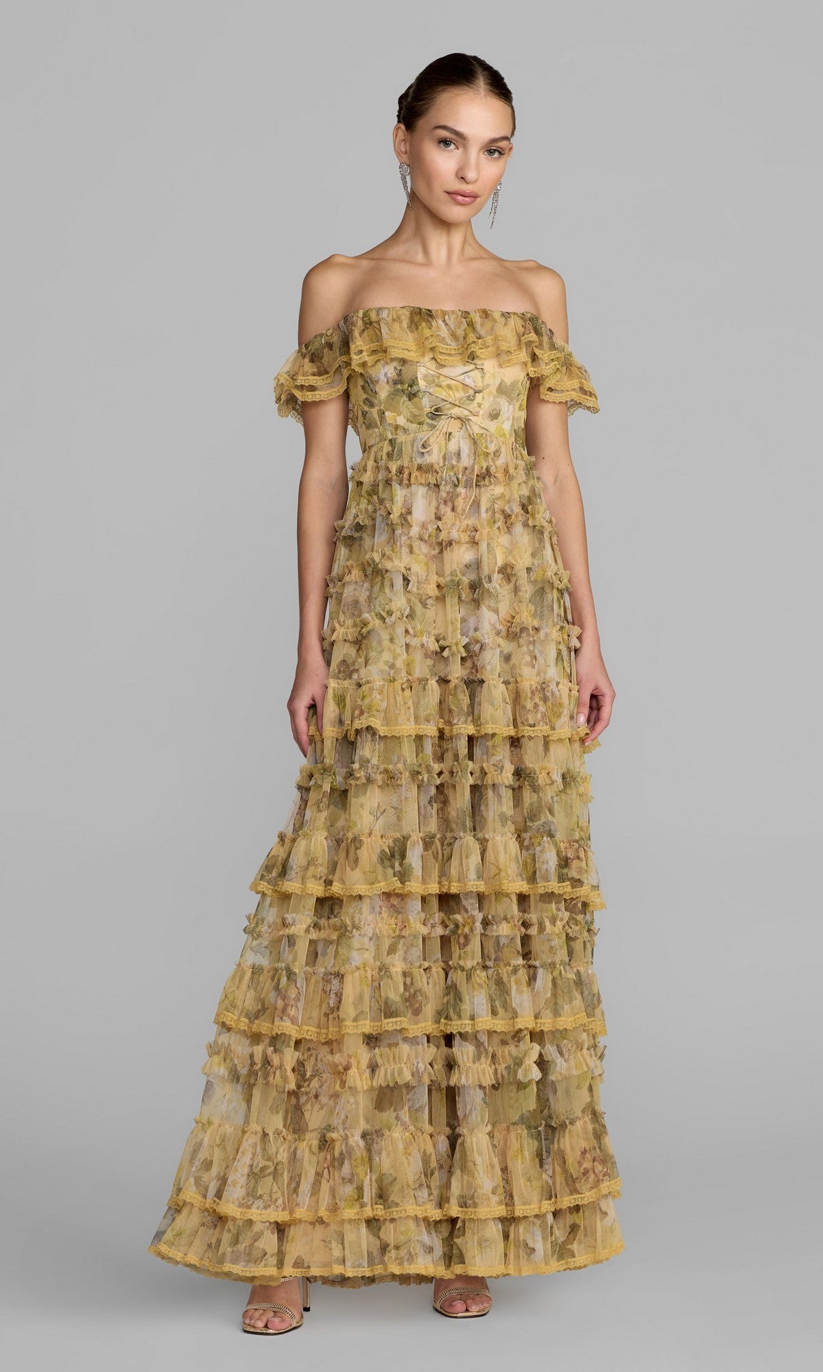 A woman wearing the Mac Duggal 8418 long formal dress in buttercream, featuring floor-length tiers and delicate ruffles with an off-the-shoulder design, stands against a plain gray background.